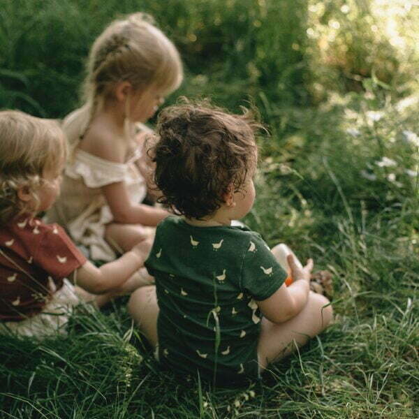 Children sitting on the meadow and a little boy is wearing a green wrapover bodysuit with short sleeves and a little geese pattern