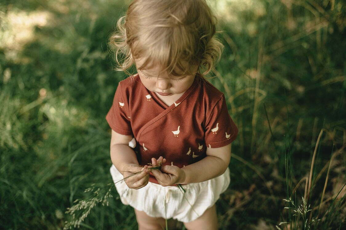 Ein Mädchen im Garten gekleidet in Baby Wickelbody aus Bio-Baumwolle mit GOTs Zertifikat und kurzen Ärmel mit Gänse Muster