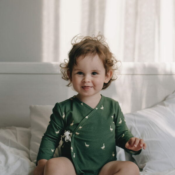 a boy on a bed wearing  Little Geese organic cotton a wrapover bodysuit with green geese pattern