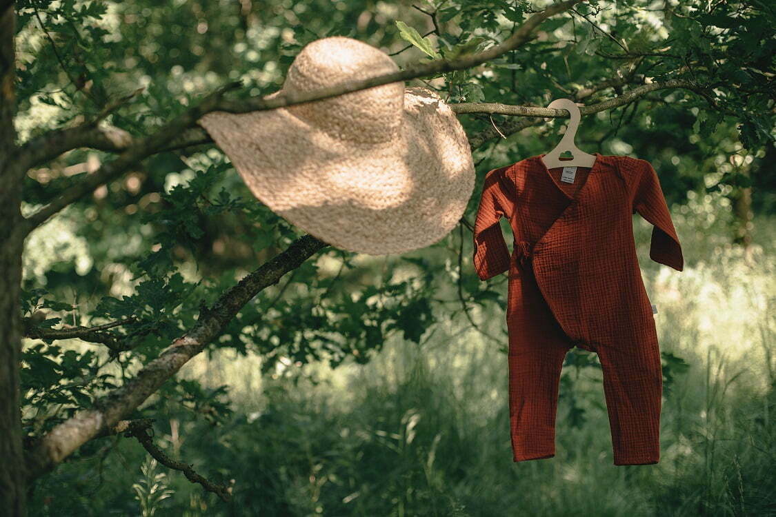 auf einem Ast hängender Baby Overall aus Musselin Bio Baumwolle in rotbrauner Farbe